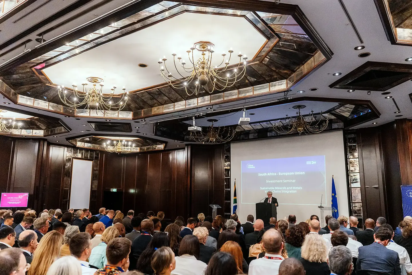 A full hotel ballroom listening to a keynote under chandeliers