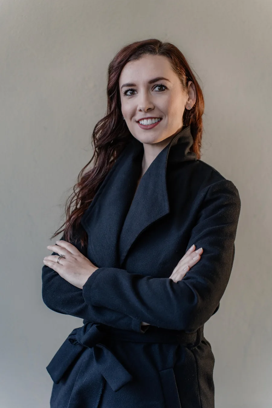 Corporate headshot of a smiling woman with long auburn hair in a black coat, arms folded, against a plain grey wall
