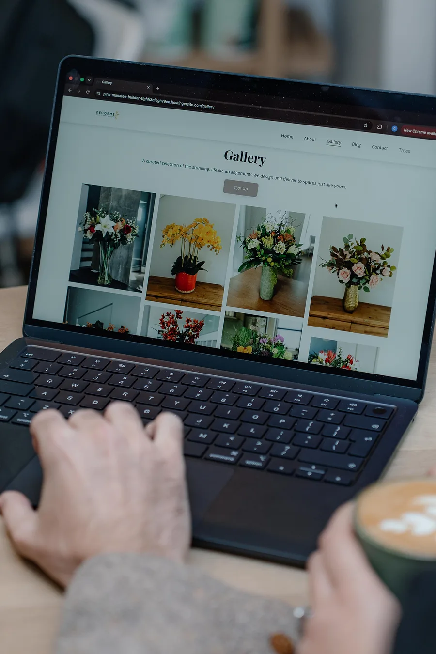 Hands on a laptop showing a florist's online gallery, with two lattes on a wooden cafe table