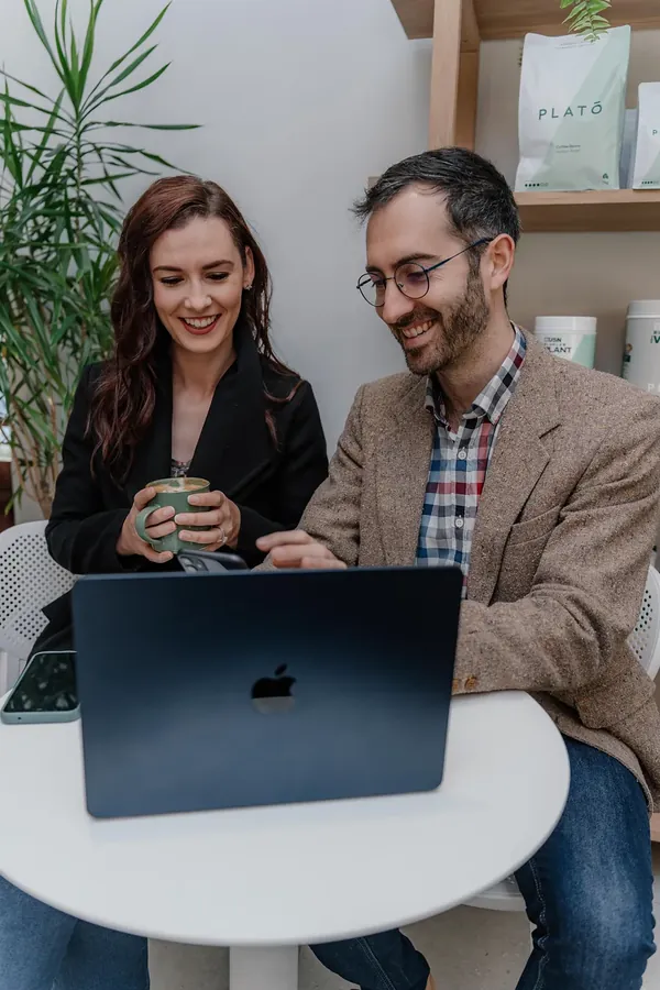 Two colleagues laughing over coffee and a laptop at a cafe