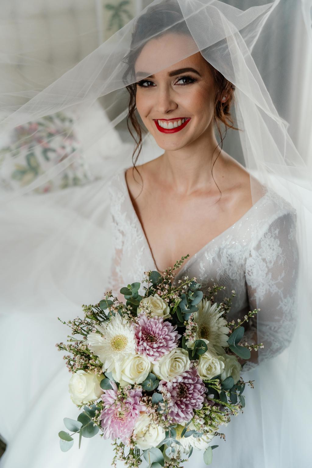 The bride smiling under her veil, holding a bouquet of white and pink flowers, Gunther and Lara's wedding