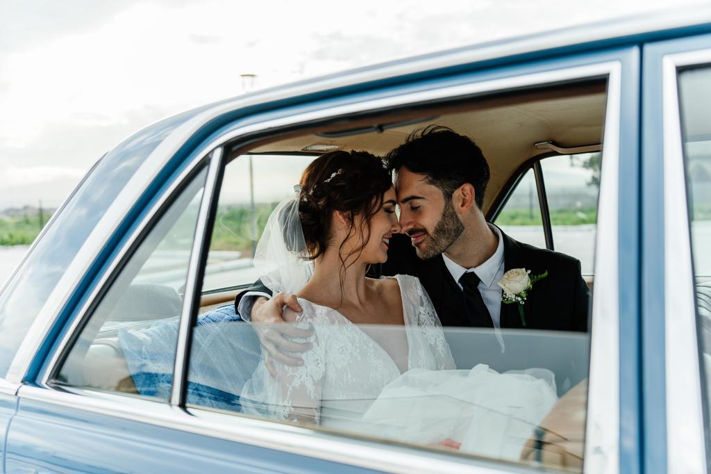 A bride and groom forehead to forehead in the back seat of a vintage blue car