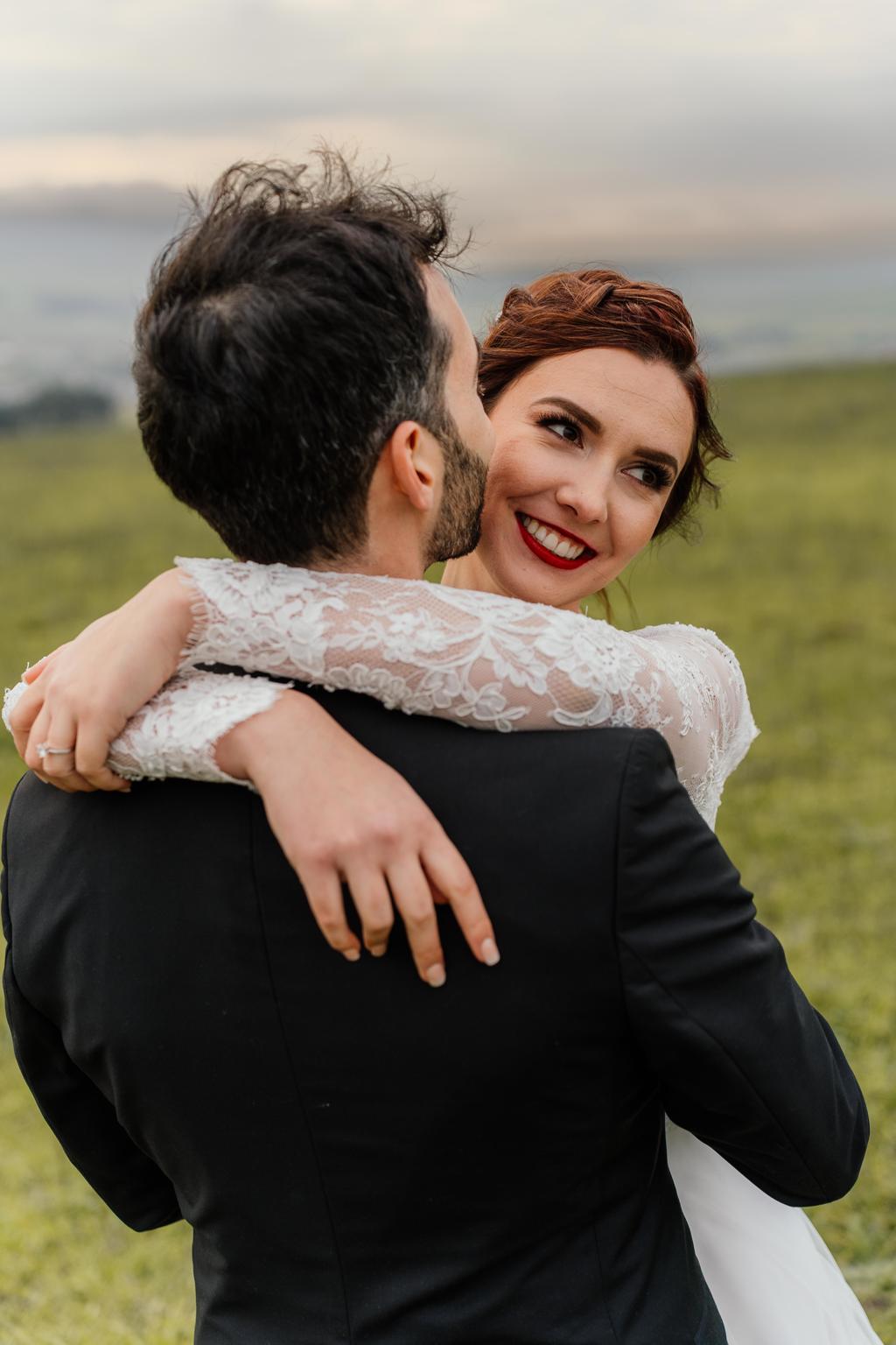 The bride smiling over the groom's shoulder as they embrace in a field, Gunther and Lara's wedding
