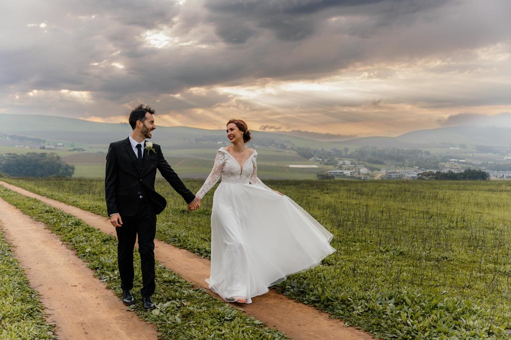 Bride and groom walking a dirt road under a wide evening sky