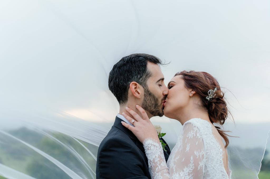 A bride and groom kissing as her veil lifts in the wind