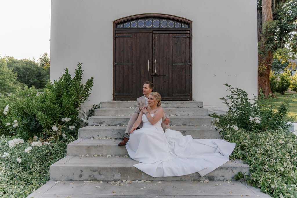 Henry and Kathrin sitting together on the stone steps of a white chapel, her dress spread across the steps among white roses