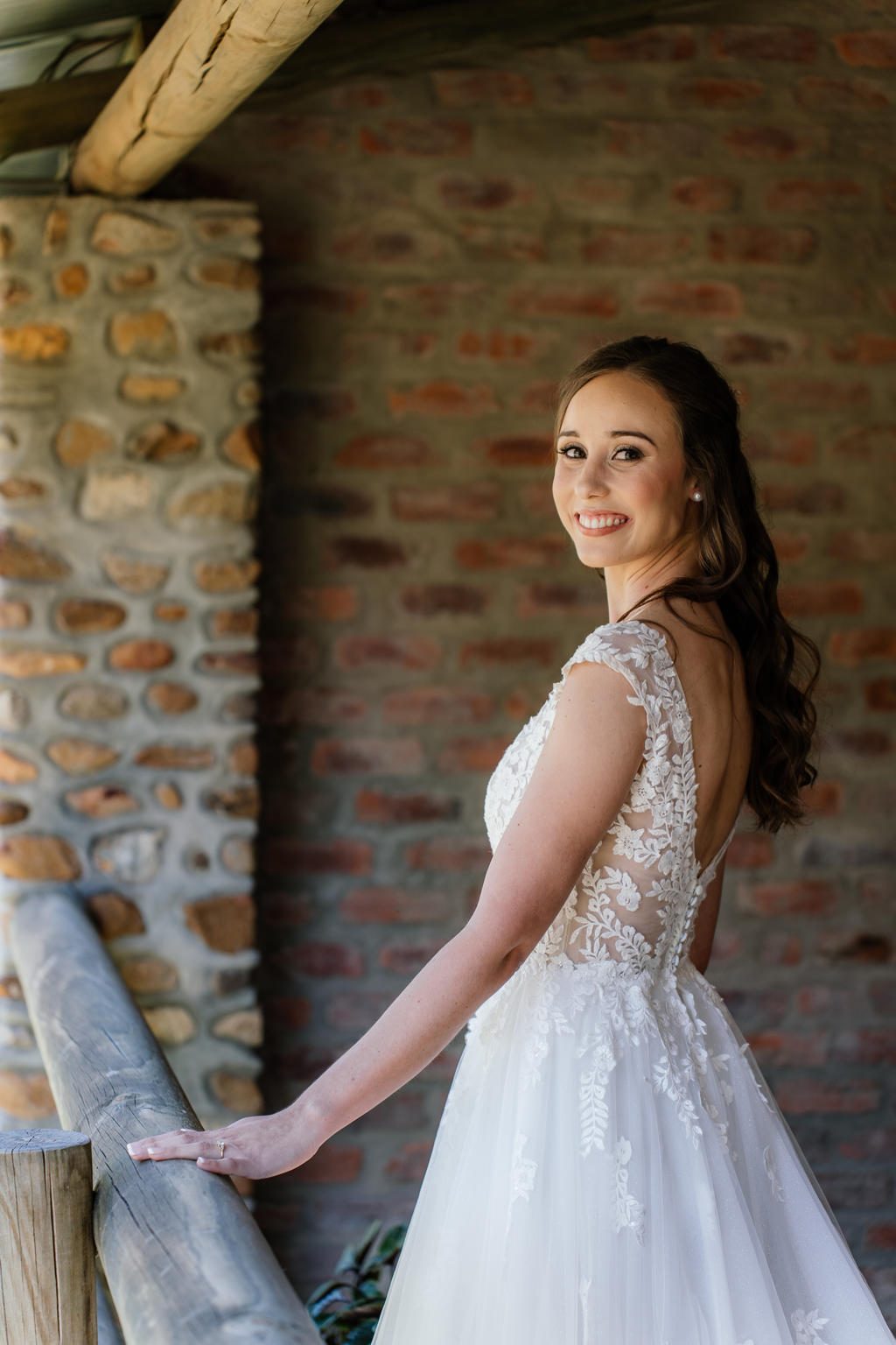 The bride in her open-back lace dress looking over her shoulder beside a brick wall, Iain and Naomi's wedding