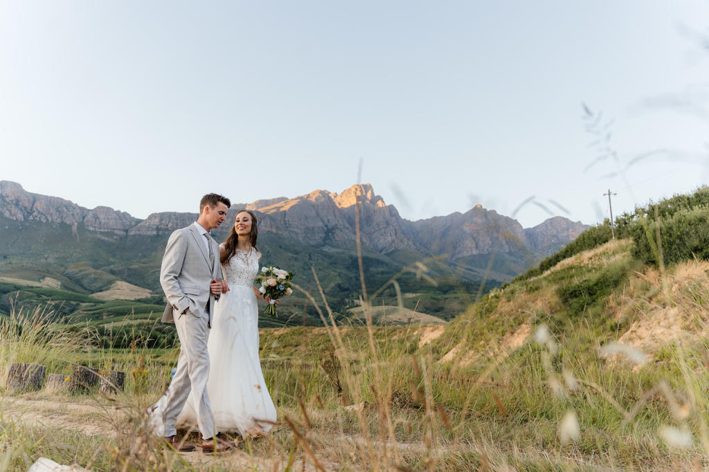 Bride and groom walking through tall grass below the mountains, Iain and Naomi's wedding