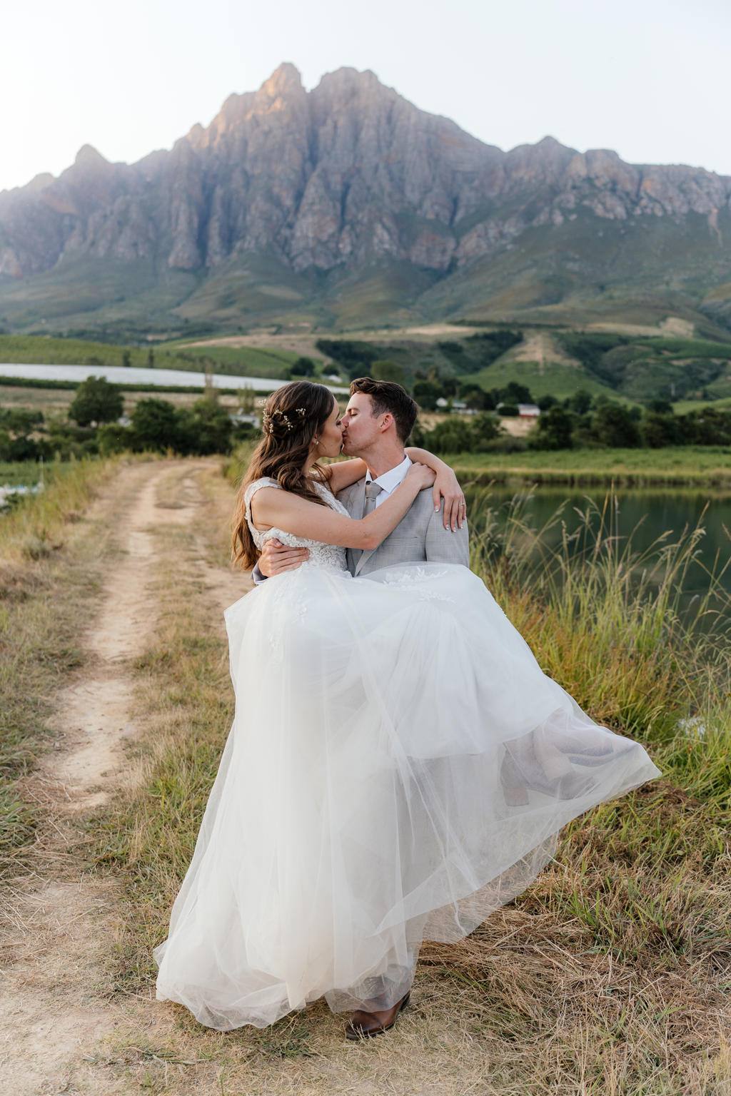 Groom carrying the bride along a farm track, kissing, below the mountains