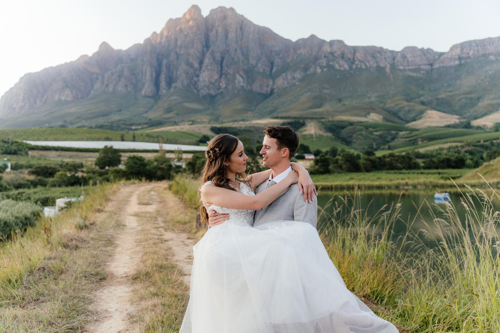 The groom holding the bride in his arms beside a farm dam below the mountains, Iain and Naomi's wedding