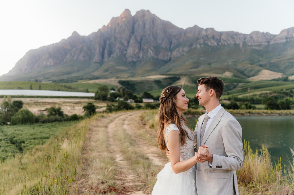 Bride and groom dancing on a farm track beside a dam, mountains behind, Iain and Naomi's wedding