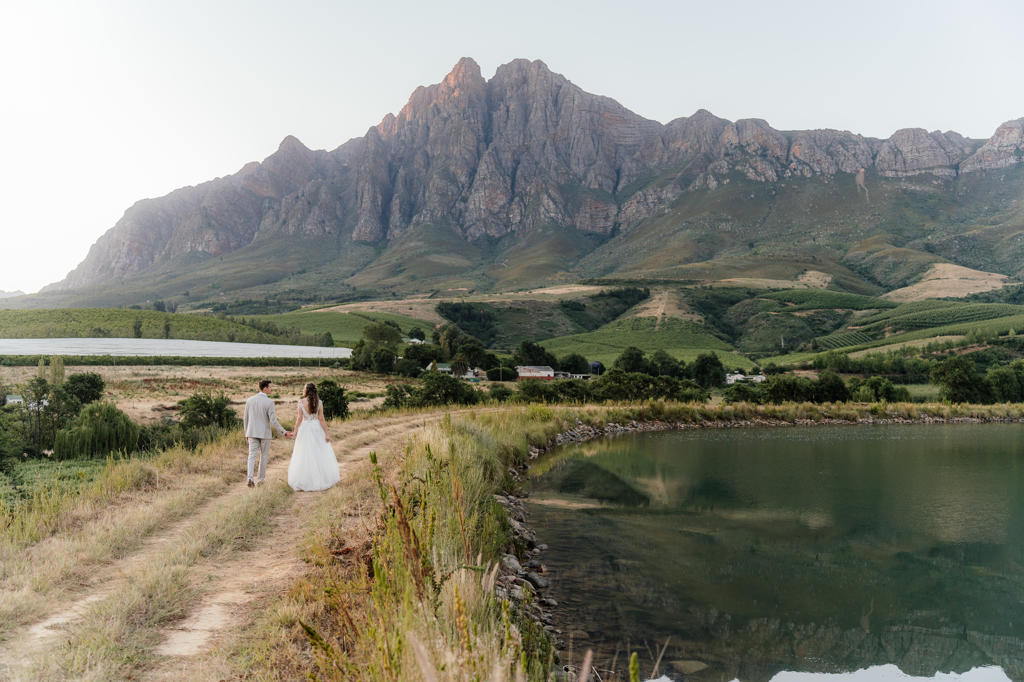 A bride and groom walking along a farm dam below the mountains