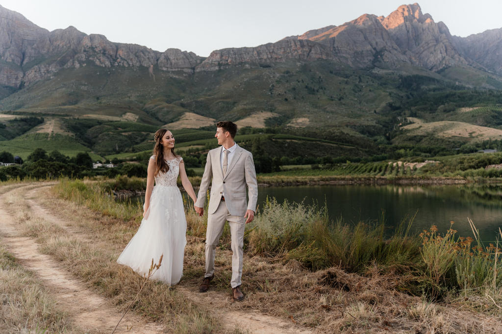 Bride and groom walking hand in hand beside a dam below the mountains, Iain and Naomi's wedding
