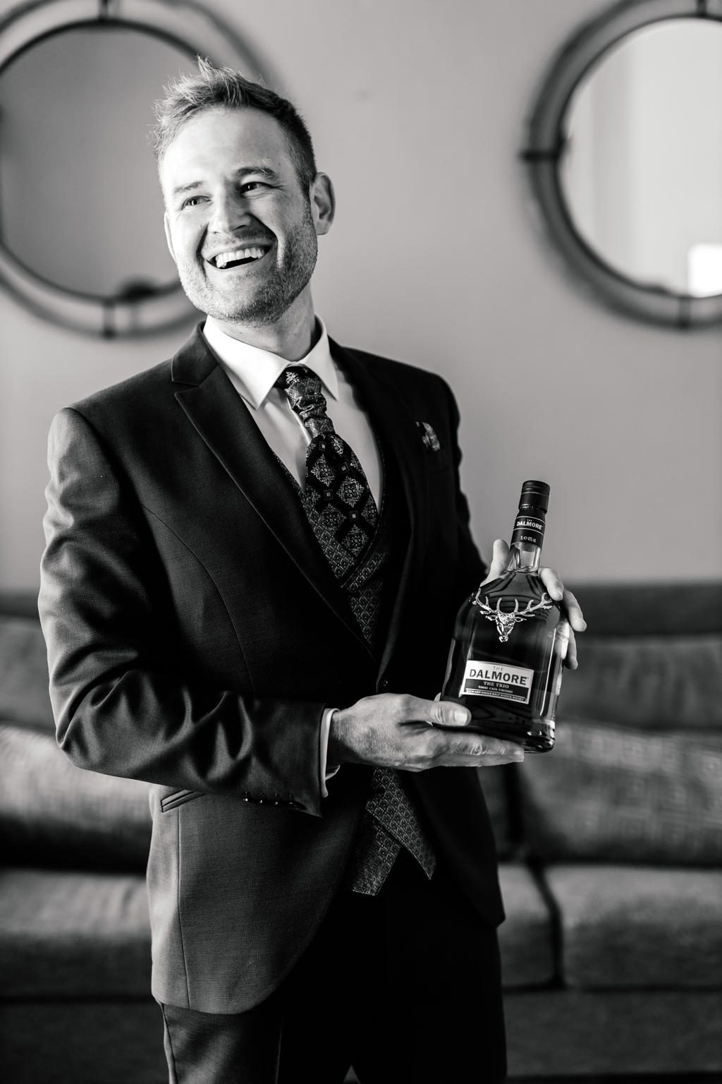 Black-and-white portrait of the groom laughing, holding a bottle of whisky, Ian and Katharina's wedding