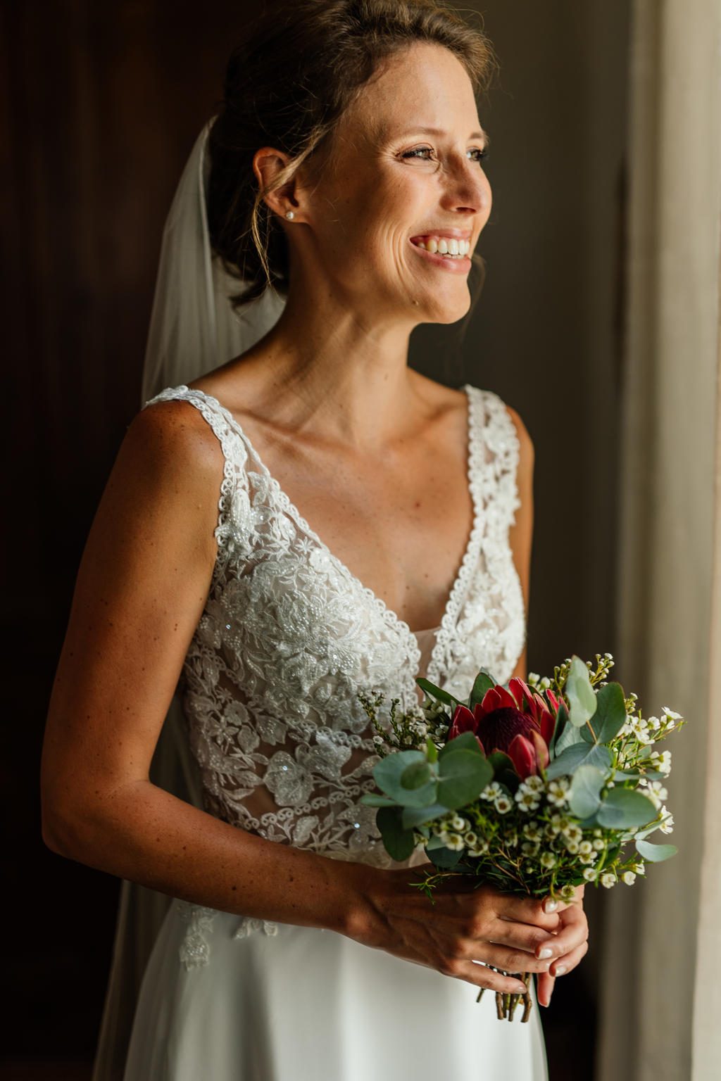 The bride smiling by a window, holding a bouquet of red and white flowers, Ian and Katharina's wedding