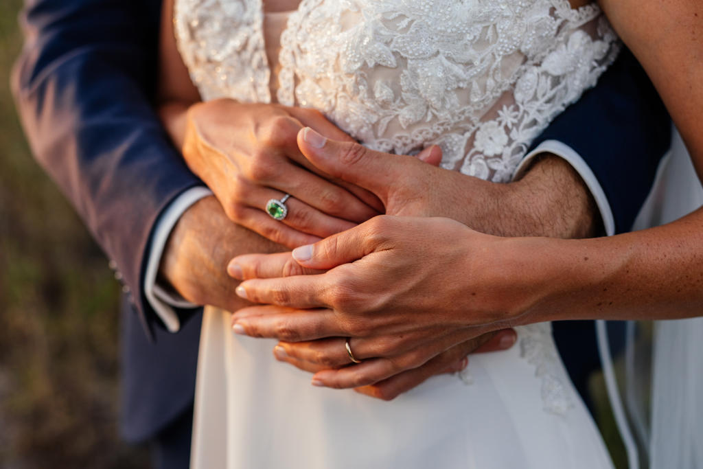 The groom holding the bride from behind, their hands and rings together over her lace dress, Ian and Katharina's wedding