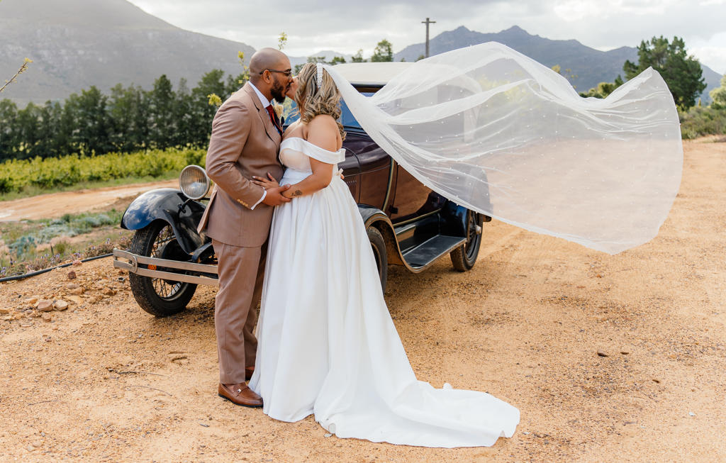 Bride and groom kissing beside a vintage car, her veil lifting in the wind
