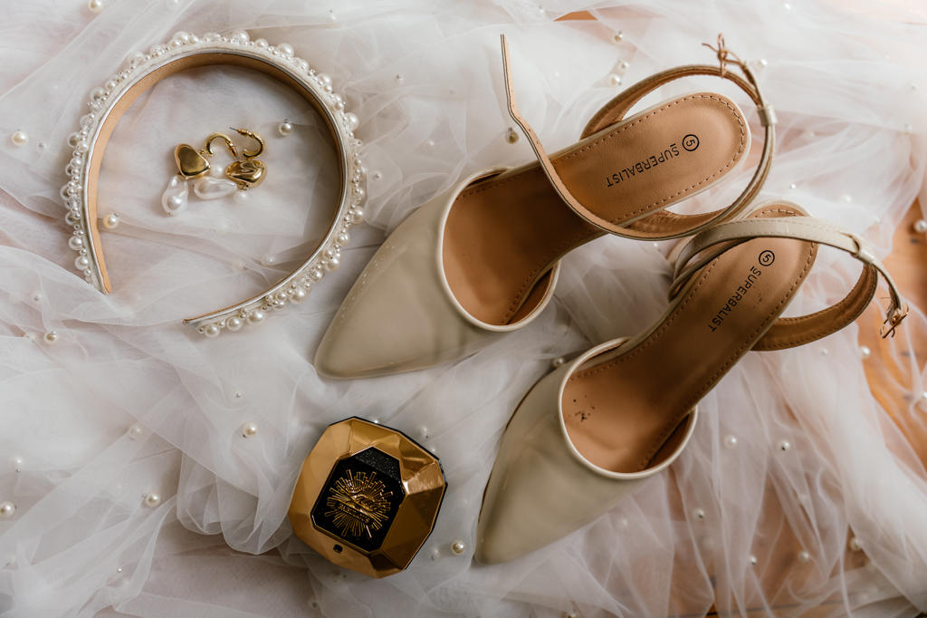 Bridal shoes, a pearl headband, pearl earrings and perfume laid out on tulle
