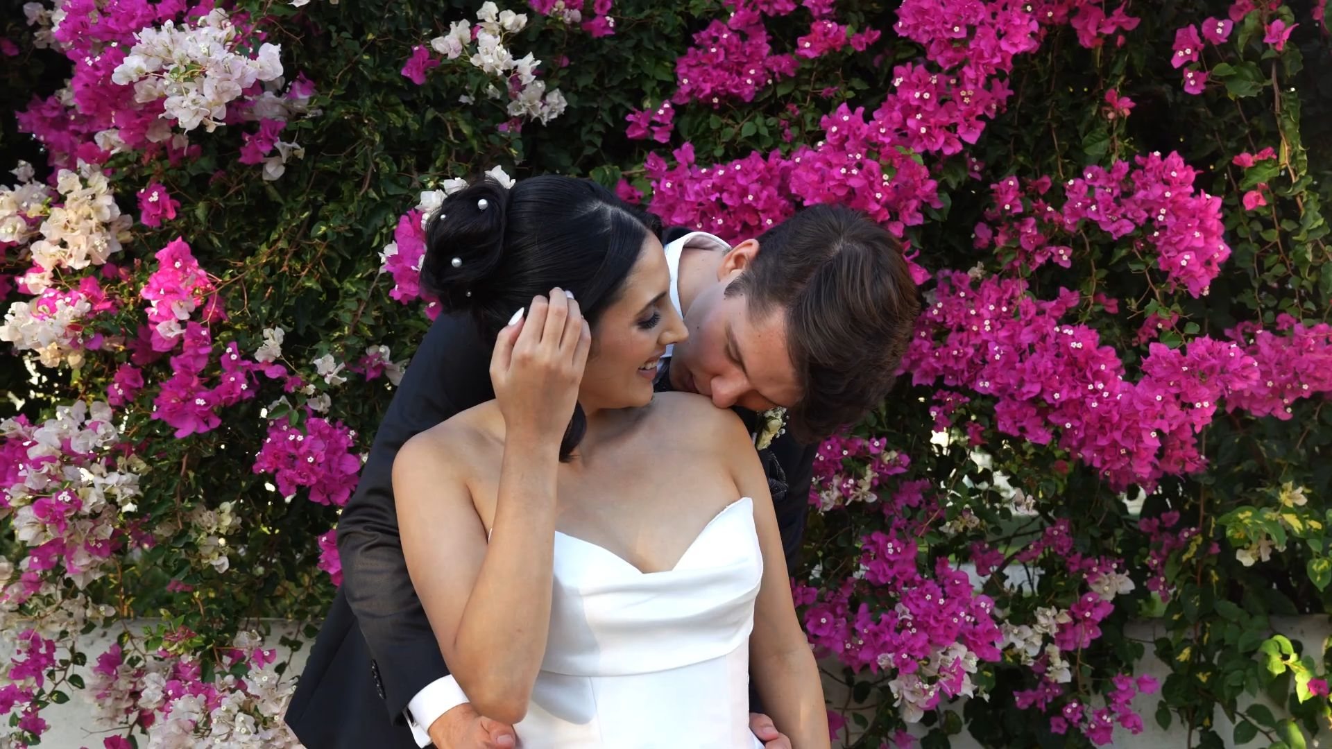 Luke kissing Leila on the shoulder in front of a wall of pink bougainvillea