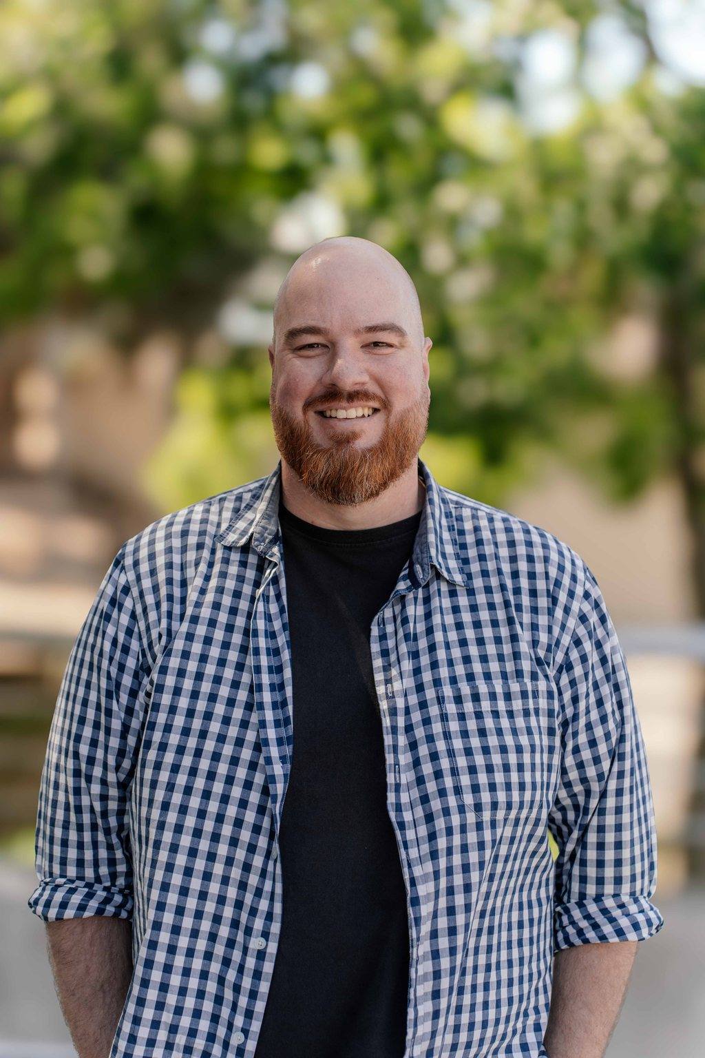 Kobus Pretorius, founder of Red Beard Photo & Film, smiling outdoors in a checked shirt under green trees