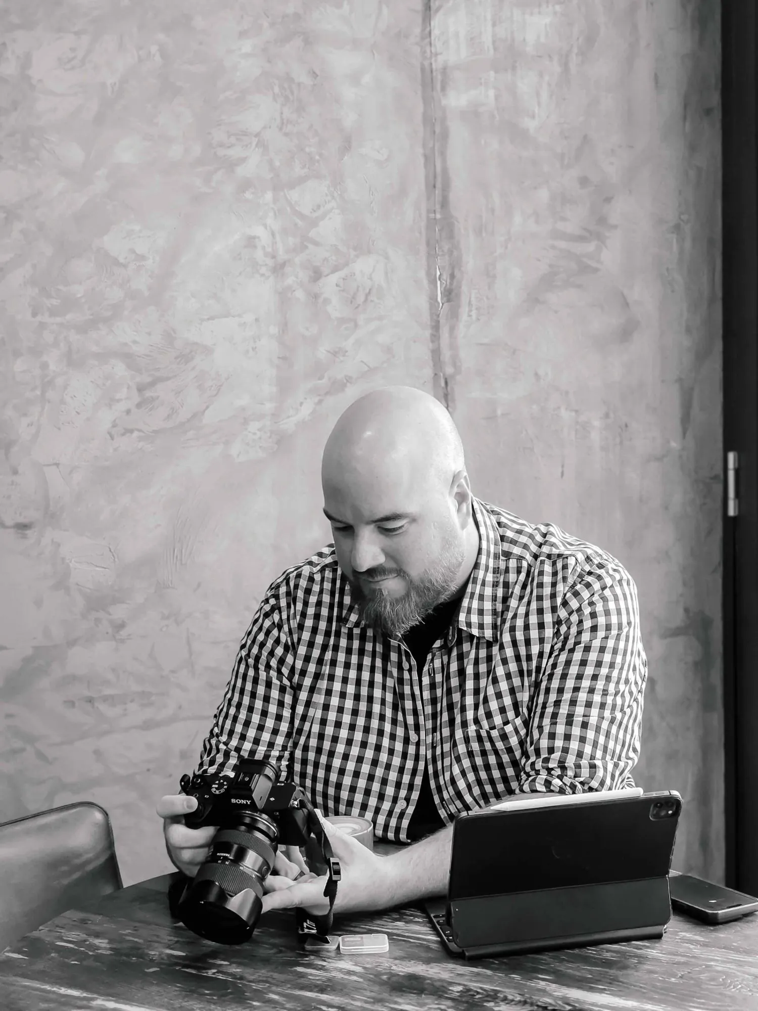 Black-and-white portrait of Kobus Pretorius at a café table, checking a shot on his camera beside his tablet
