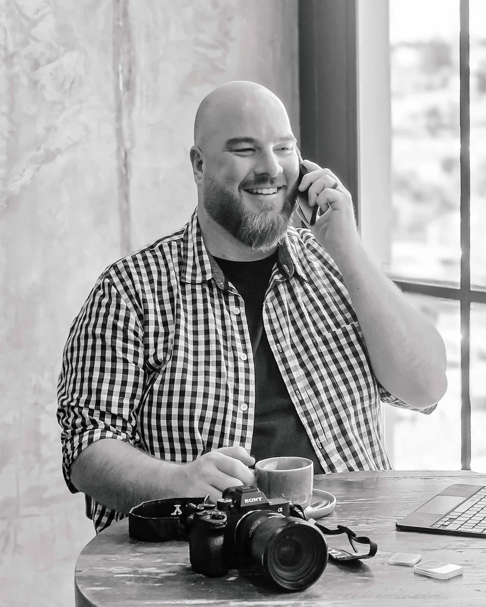 Kobus smiling on a phone call at a cafe table, his camera and a coffee in front of him