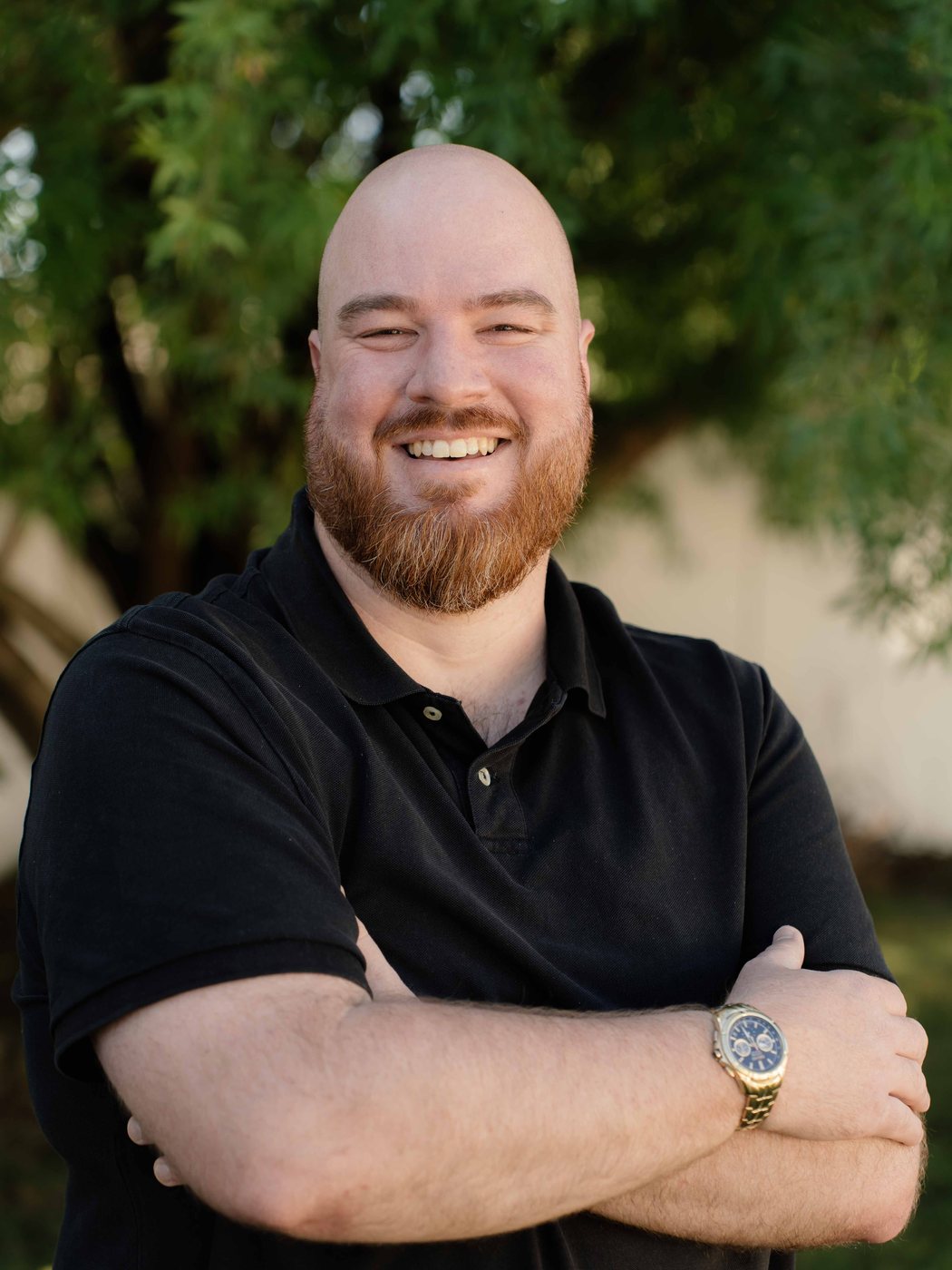 Kobus Pretorius smiling with his arms folded, in a black shirt in front of a leafy tree