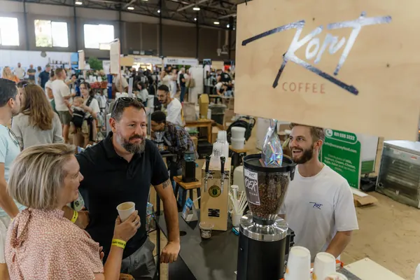 The barista smiling behind the grinder as a customer looks on, Kort Coffee