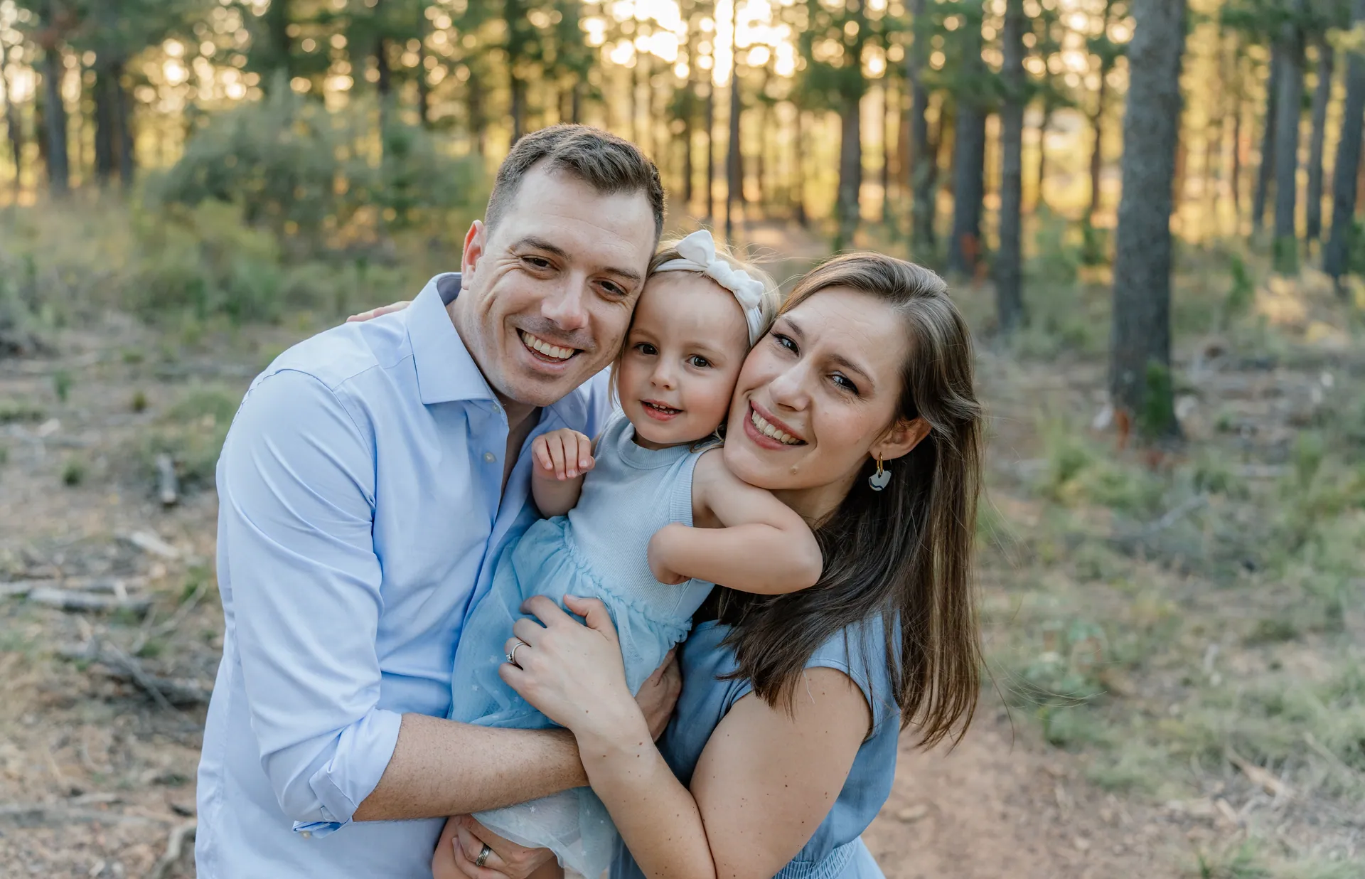 A mother and father holding their little girl between them in golden light among the pines, the May family shoot
