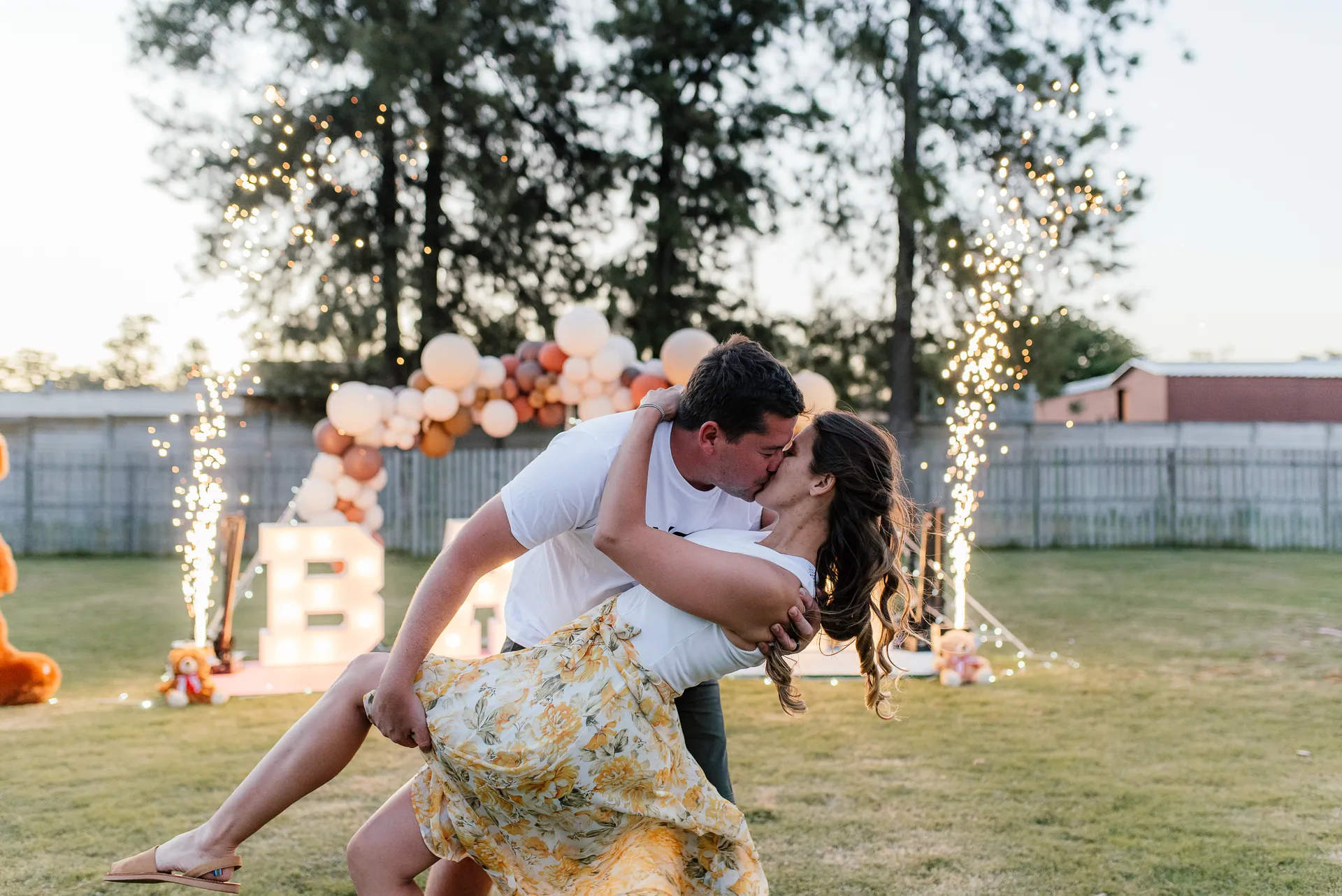 A couple sharing a dip kiss in front of light-up BABA letters and sparklers at their gender reveal