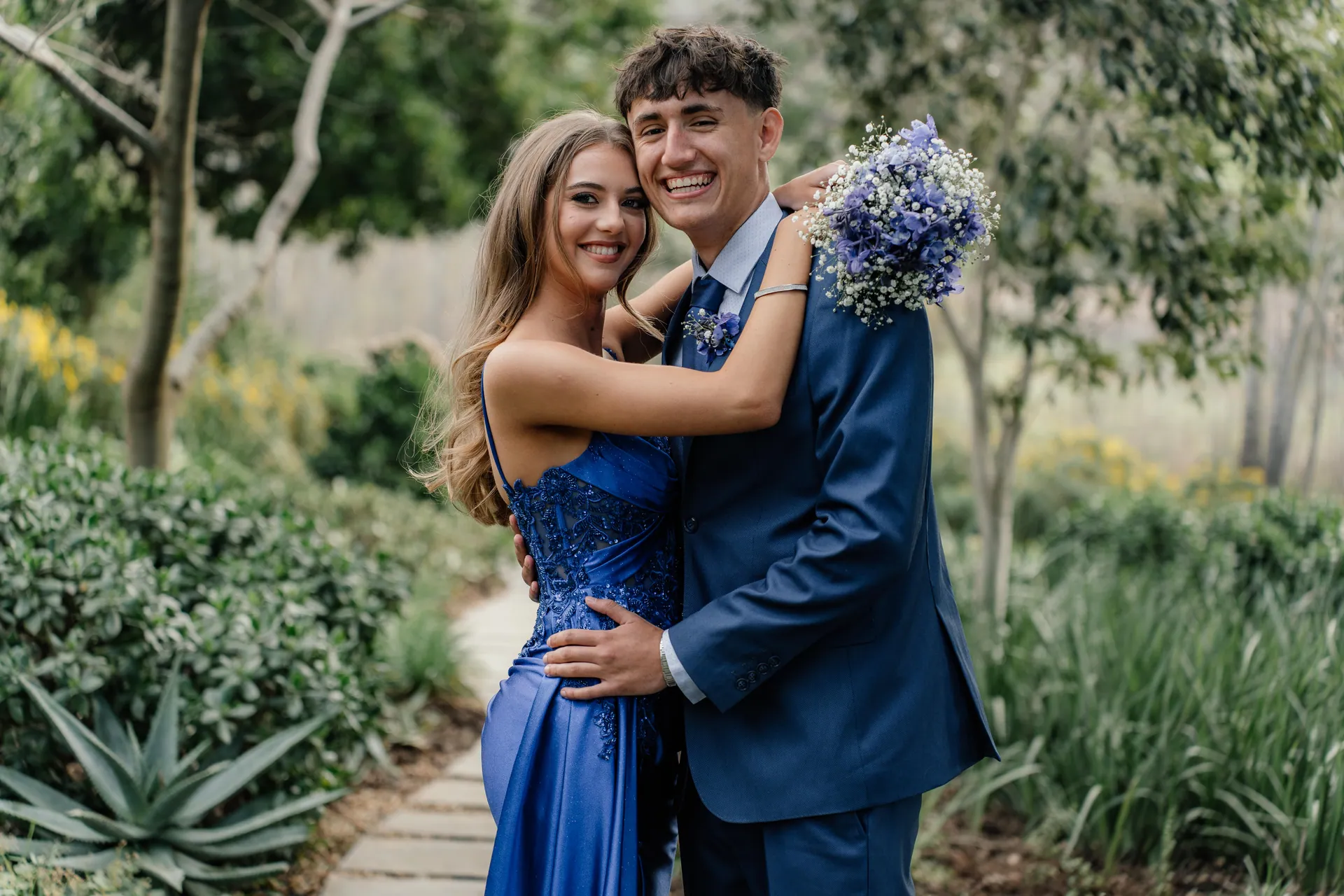 A matric farewell couple in a blue gown and navy suit laughing together in a garden, Jacinda