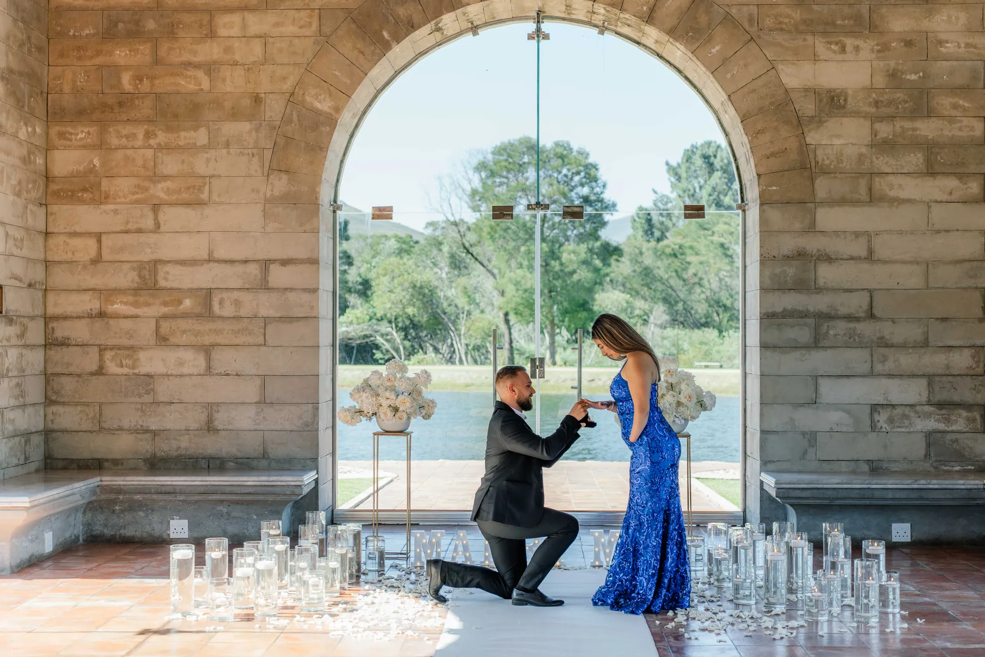 A man on one knee proposing beneath a stone archway overlooking a lake, Jonathan and Matilda