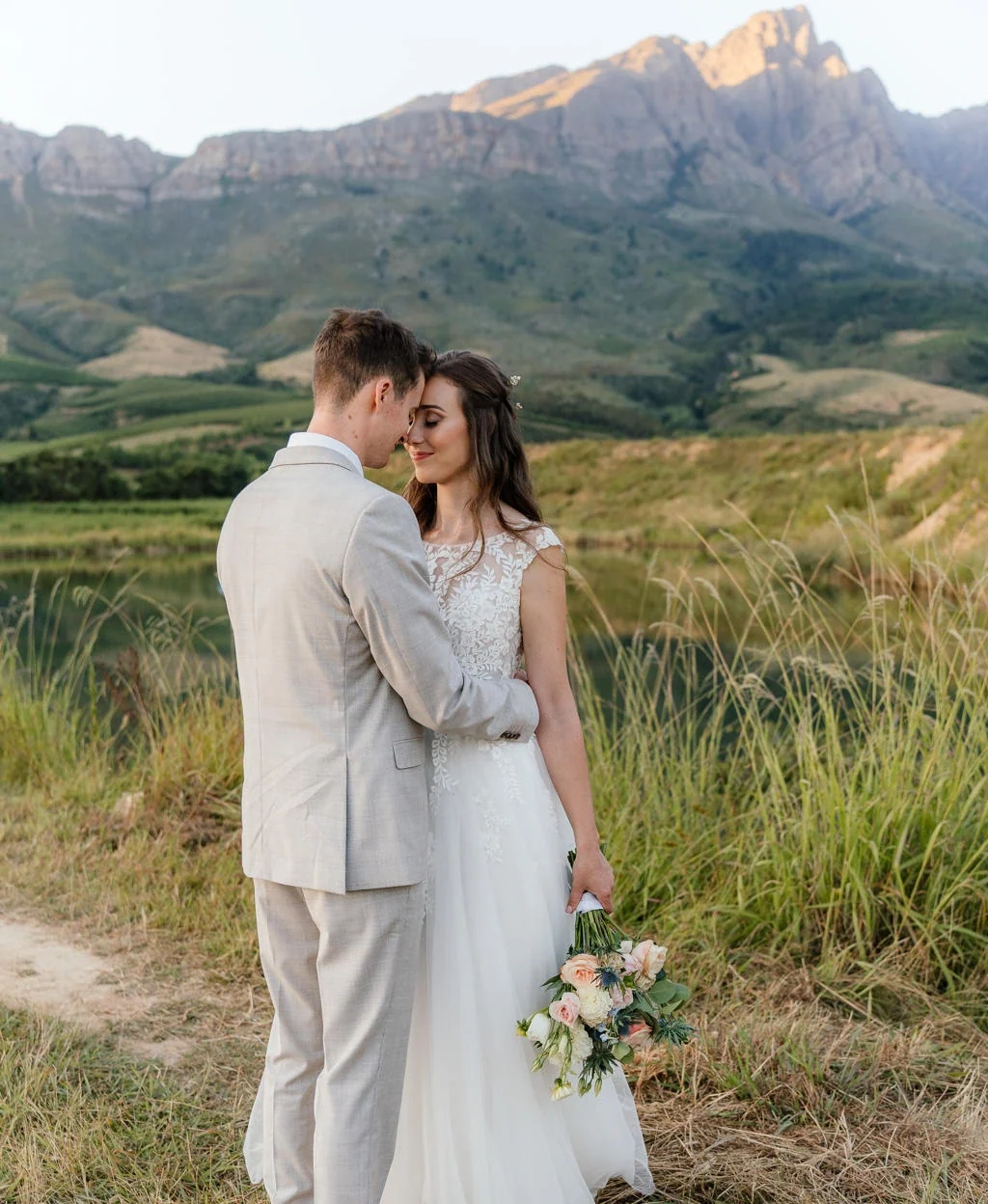Bride and groom standing close in a field below the Boland mountains at sunset